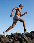 Man wearing Flow shorts running over rocky terrain against a clear blue sky, showcasing active movement and style.