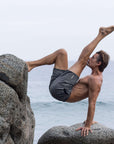 Man performing a yoga pose on rocks by the ocean while wearing 4-way stretch swerve shorts in heather grey.