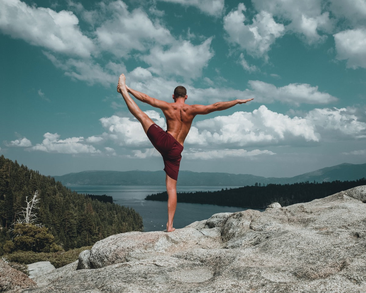 Man practicing flexibility on a rocky outcrop with a scenic lake and mountains in the background.